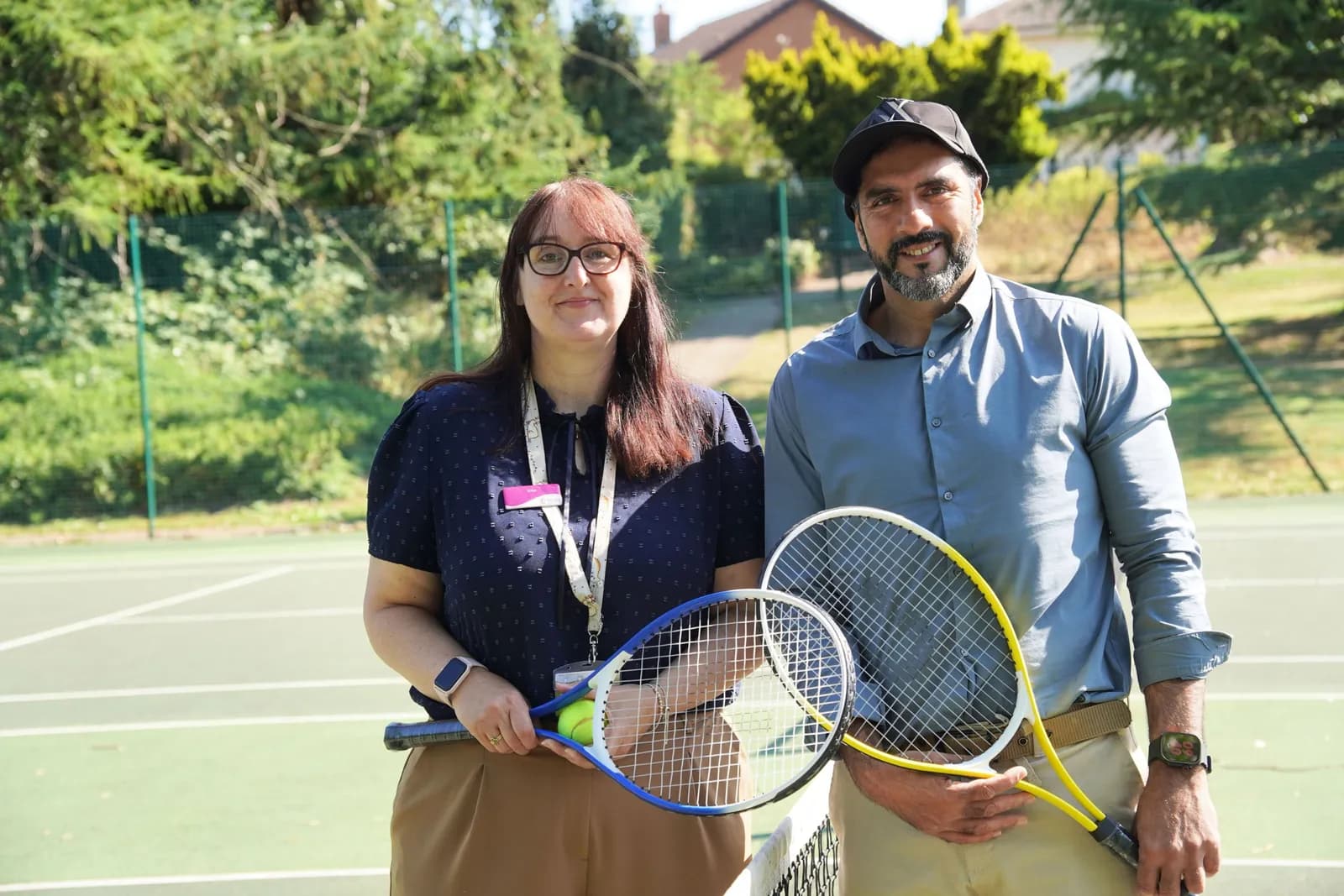 A man and a woman standing on a tennis court, both holding tennis rackets, smiling at the camera on a sunny day.